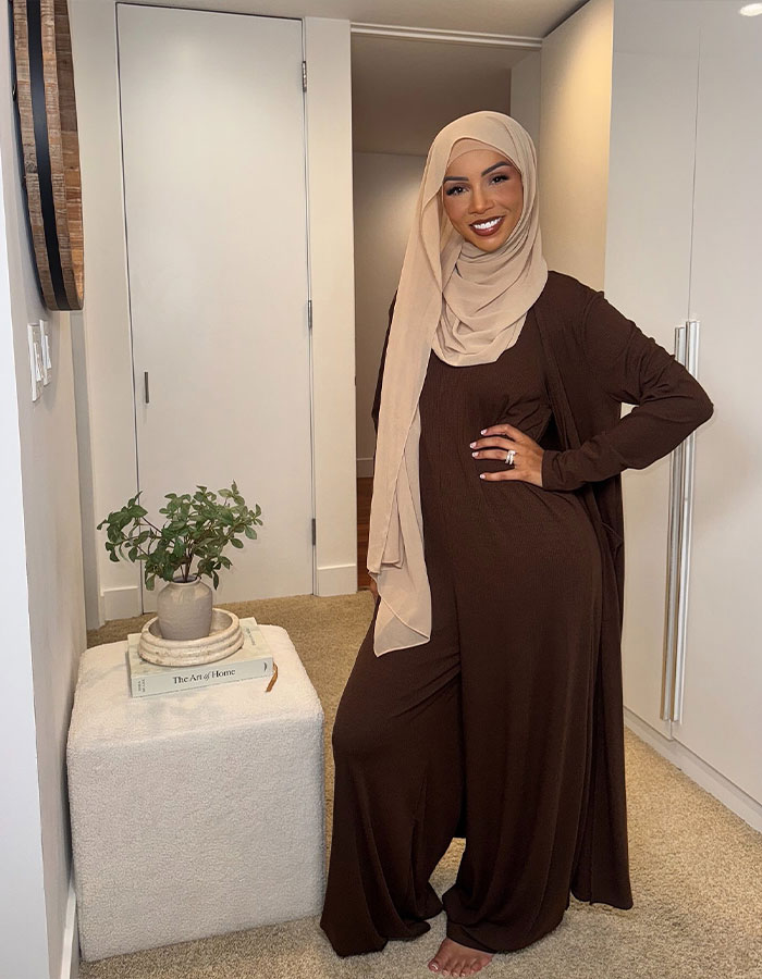 Influencer wearing a beige hijab and brown modest outfit, smiling and posing indoors near a small table with plants. Influencer wearing a beige hijab and brown modest outfit, smiling and posing indoors near a small table with plants.