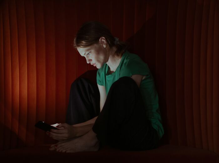 Young woman sitting alone in dim light, looking at phone, depicting emotions related to cheating red flags in relationships. - 28