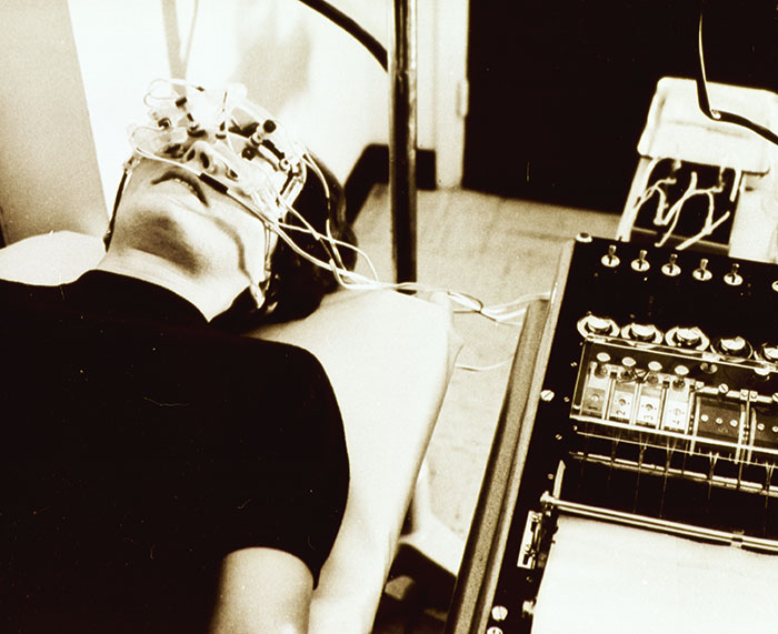 Patient lying down connected to unsettling medical devices from the past with wires and monitoring equipment attached.