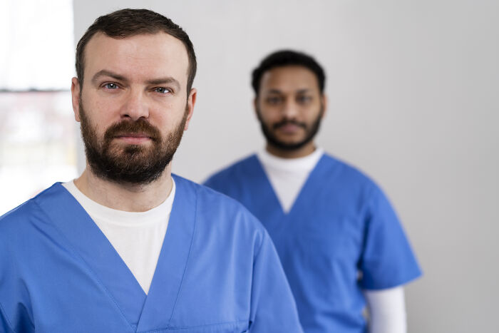 Two doctors in blue scrubs, one looking serious in forefront, representing stories of impostor patients and medical insights.