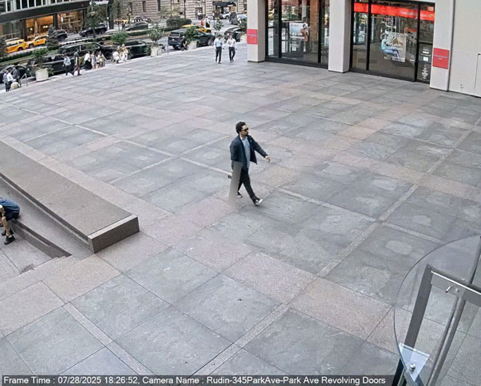Man walking alone on a large plaza outside a building with revolving doors in a busy urban area.