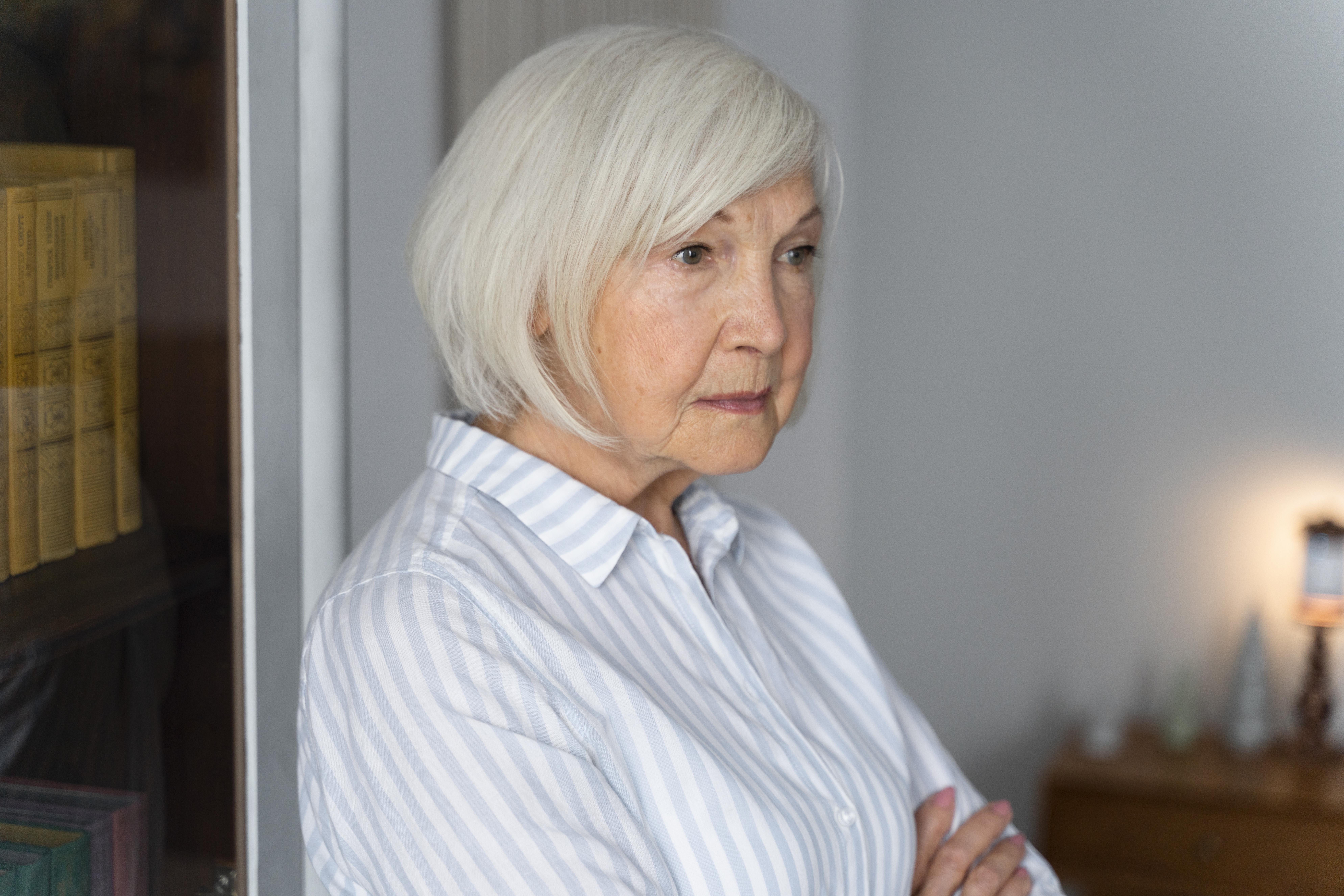 Elderly woman with white hair standing pensively indoors highlighting family drama over inherited house.