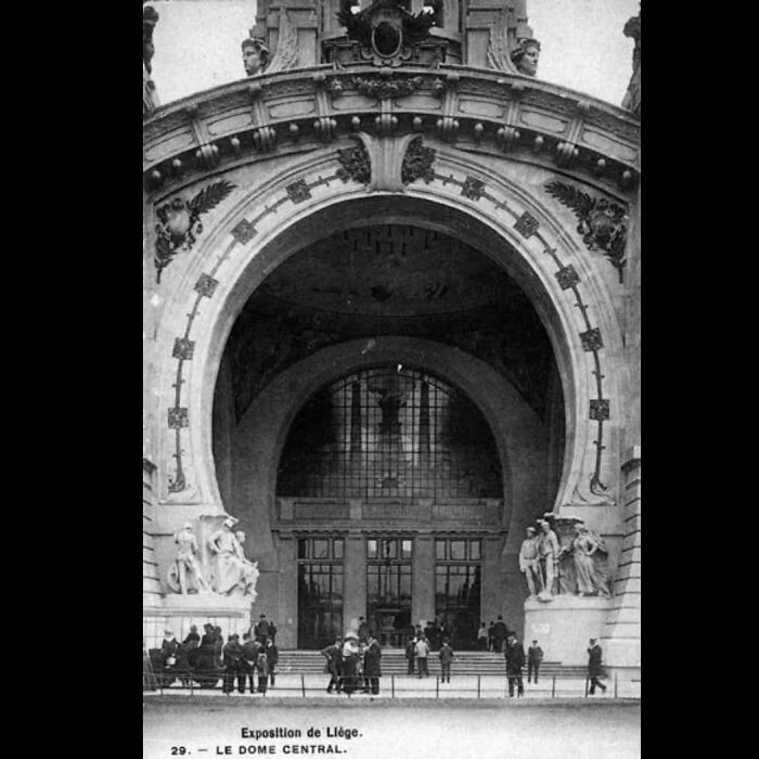 Black and white photo of old architecture featuring a grand arched entrance and detailed sculptures with people gathered outside.