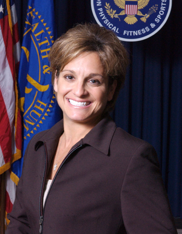 Olympic gold medalist Mary Lou Retton smiling in a professional setting with US flags and an official seal behind her