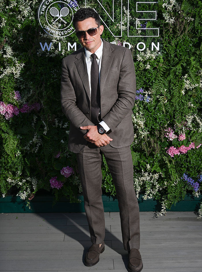 Man wearing a brown suit and sunglasses posing at Wimbledon 2025 with a floral backdrop and event logo behind him.