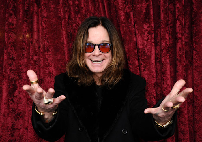 Ozzy Osbourne in black coat and red sunglasses, smiling with arms extended against a red velvet curtain background. Ozzy Osbourne in black coat and red sunglasses, smiling with arms extended against a red velvet curtain background.