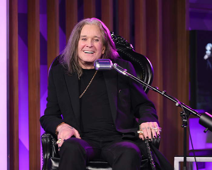 Rock God Ozzy Osbourne smiling while seated on stage with microphone, wearing black outfit and gold chain necklace. - 2