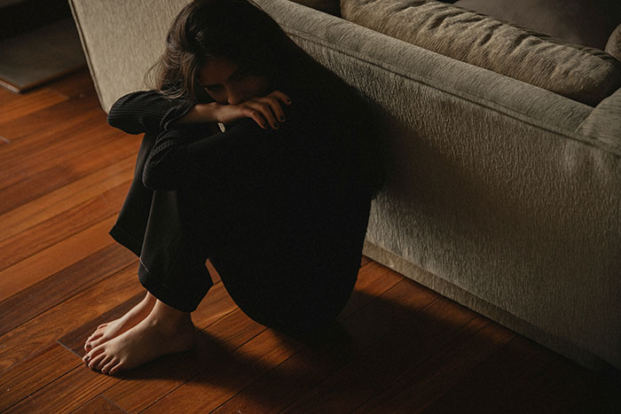 Woman sitting on wooden floor in dark clothing looking down, conveying emotion after man publicly demoted to friend by girlfriend