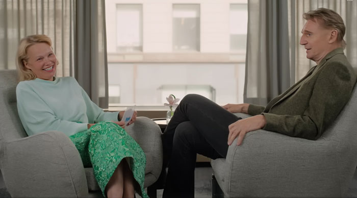 Pamela Anderson and Liam Neeson sitting in armchairs, smiling and engaged in a relaxed conversation indoors.