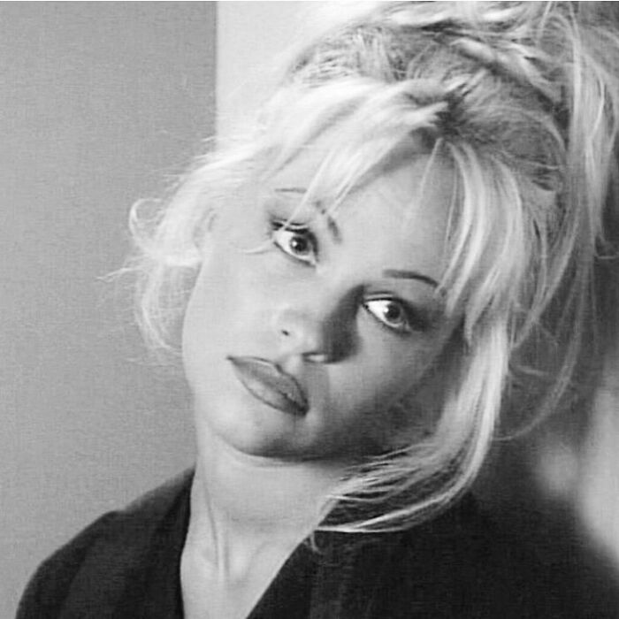 Black and white close-up of Pamela Anderson looking thoughtful with tousled hair and soft lighting in a portrait style image.