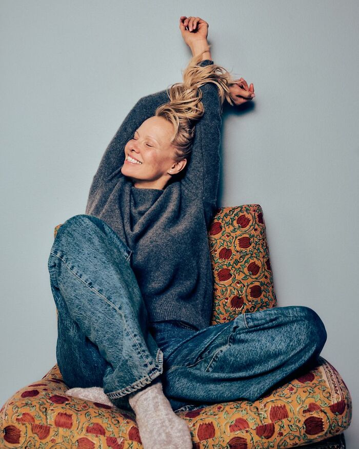 Smiling woman in casual sweater and jeans stretching while sitting on a patterned chair, reflecting Pamela Anderson dating Liam Neeson.