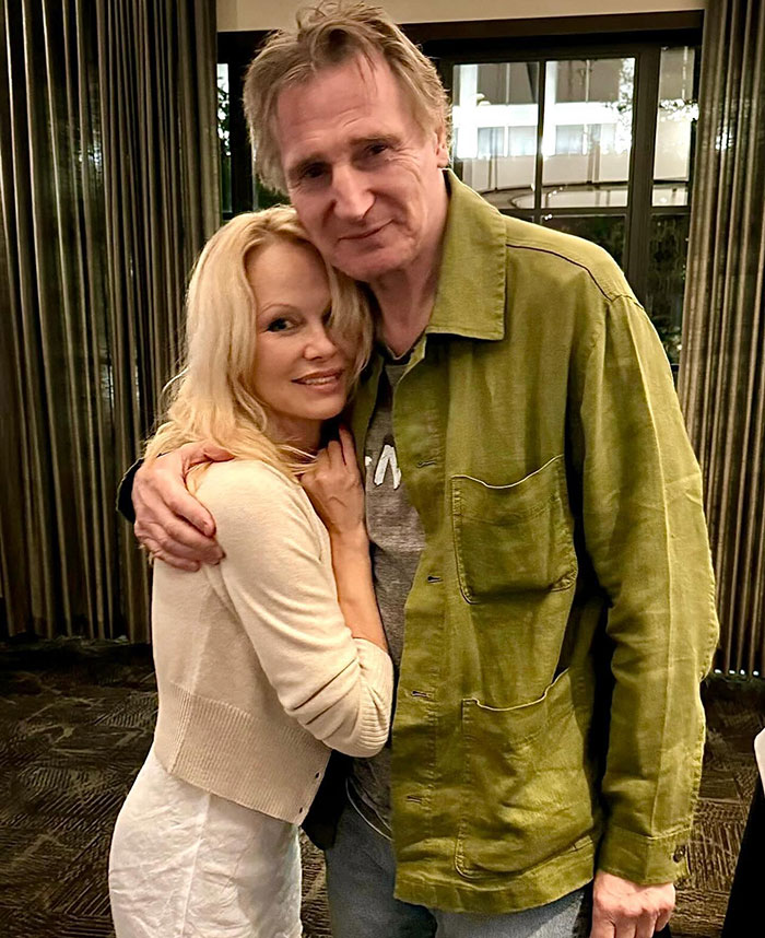 Pamela Anderson and Liam Neeson embracing warmly indoors, showing affection and closeness in a casual setting.