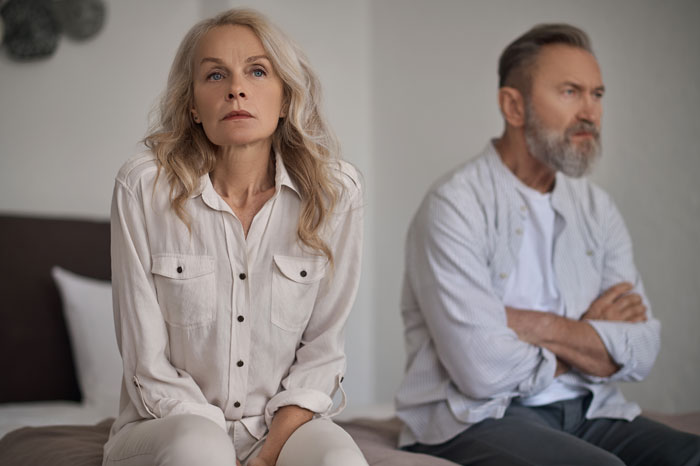 Middle-aged parents sitting apart on a bed, looking upset and distant, highlighting challenges in kid care and parenting.
