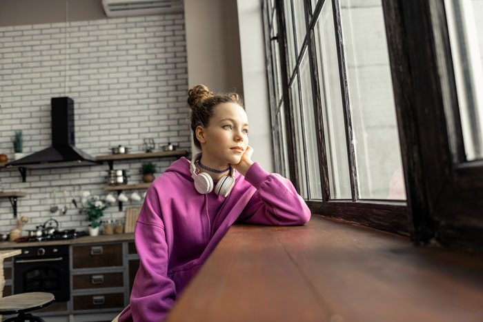 Young woman in a purple hoodie with headphones, sitting by a window, reflecting on leaving toxic parents and shared roof struggles. Young woman in a purple hoodie with headphones, sitting by a window, reflecting on leaving toxic parents and shared roof struggles.