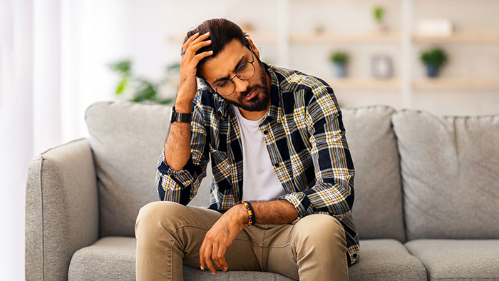 Young man looking distressed on a couch, illustrating feelings of being treated unfairly by parents favoring adopted kids. - 1