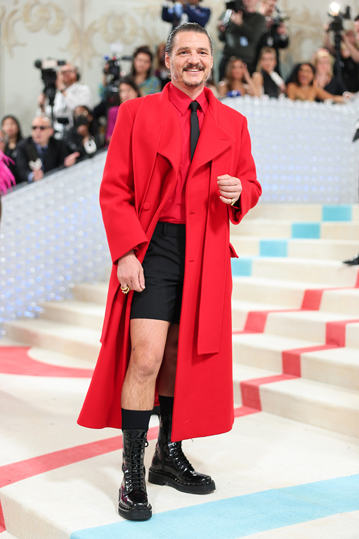 Pedro Pascal wearing a bold red coat and black boots at a high-profile fashion event with photographers in the background
