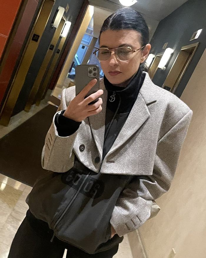 Woman wearing glasses and a grey blazer taking a mirror selfie, related to Pedro Pascal cutting ties with stylist controversy.