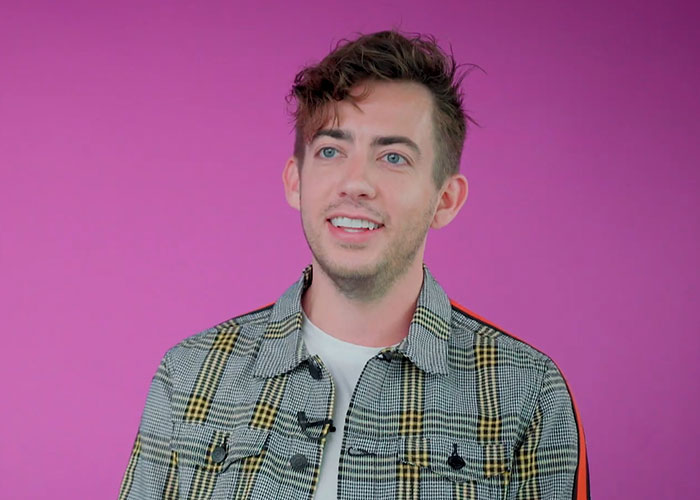 Young man sharing school memories with celebrities, wearing a checkered jacket against a pink background.