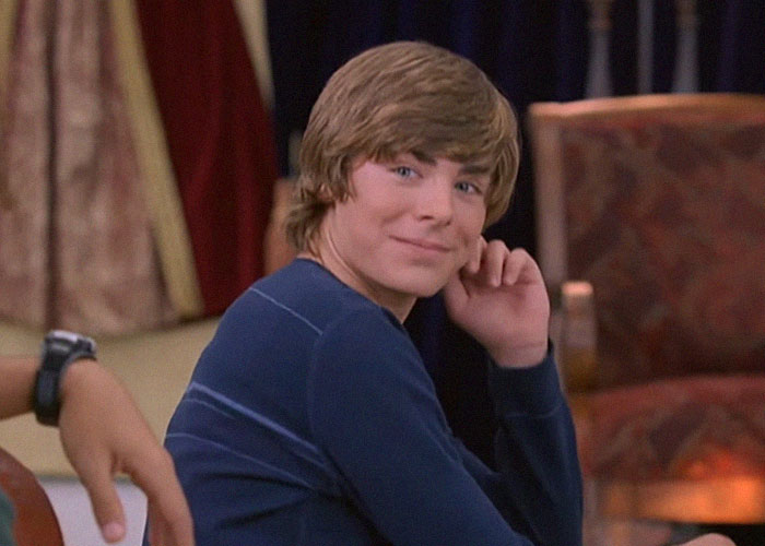 Young male celebrity sitting indoors, smiling and looking over shoulder, sharing school experiences with celebrities.