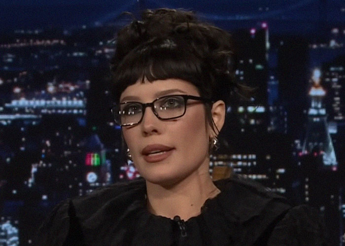 Person wearing glasses and black outfit speaking on a nighttime talk show about school with celebrities experience.