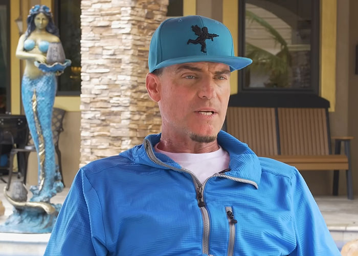 Man in blue jacket and cap sitting outdoors near a statue, sharing stories about people who went to school with celebrities.