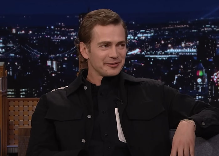Man in black jacket sitting and smiling during a nighttime talk show about people who went to school with celebrities.