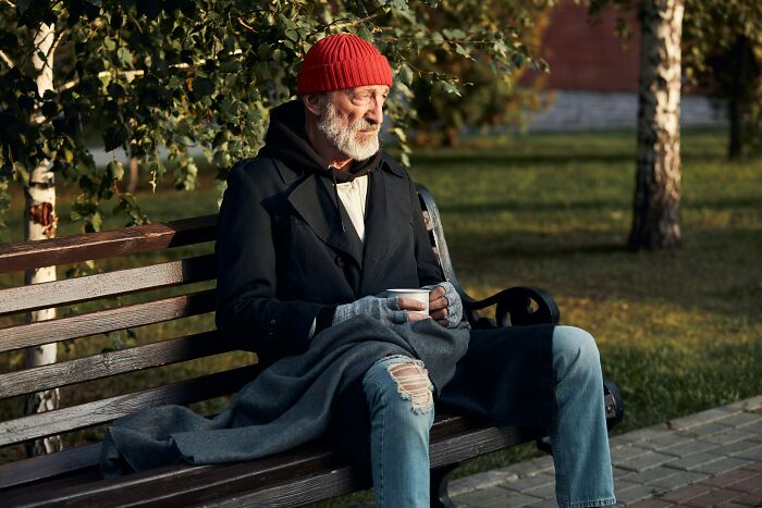 Elderly man sitting on a bench outdoors, wearing a red beanie and holding a cup, reflecting on gut feeling regrets.