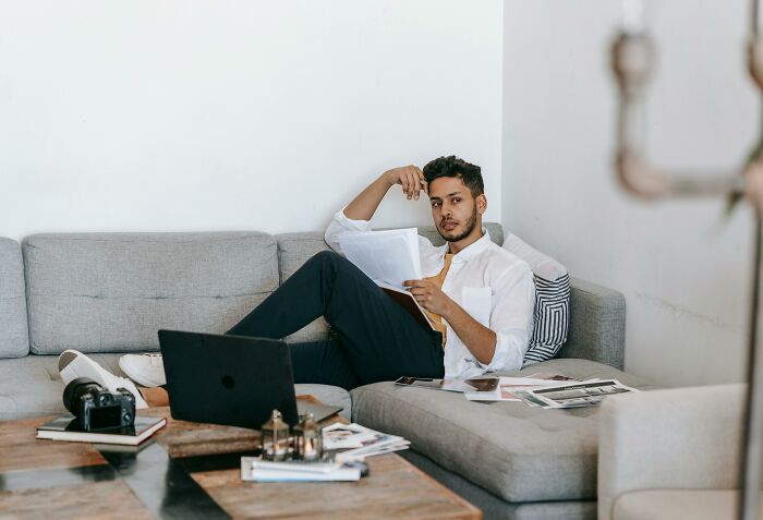 Young man reading papers on a couch with laptop and camera nearby, illustrating psychology experiments study session.