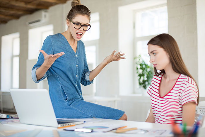 Young woman calling out her boss in an office setting while colleague looks concerned during a work confrontation.