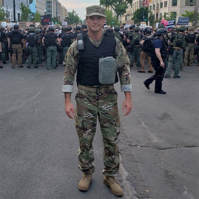 Pete Hegseth in military uniform standing on a street with law enforcement and protesters in the background.
