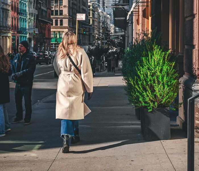 Woman walking on city sidewalk in beige coat and blue jeans, representing daily wins from feminists in urban setting.