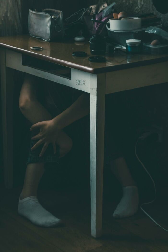 Person hiding under a table in a dimly lit room illustrating survival tips that won't help in real situations.