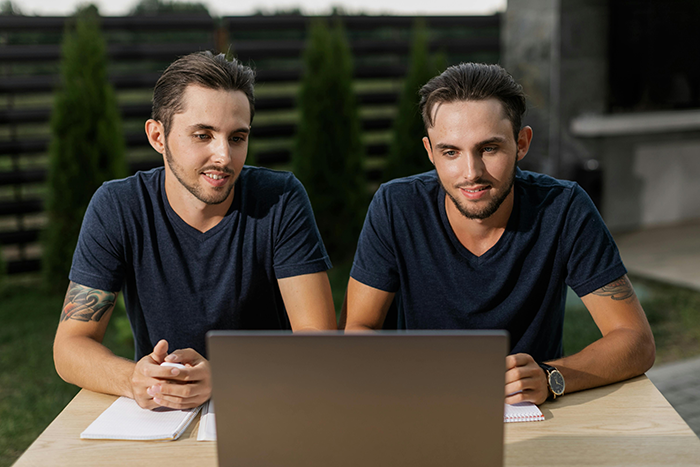 Twin gay men sitting outdoors at a table with a laptop, wearing navy shirts, discussing plans with partner.