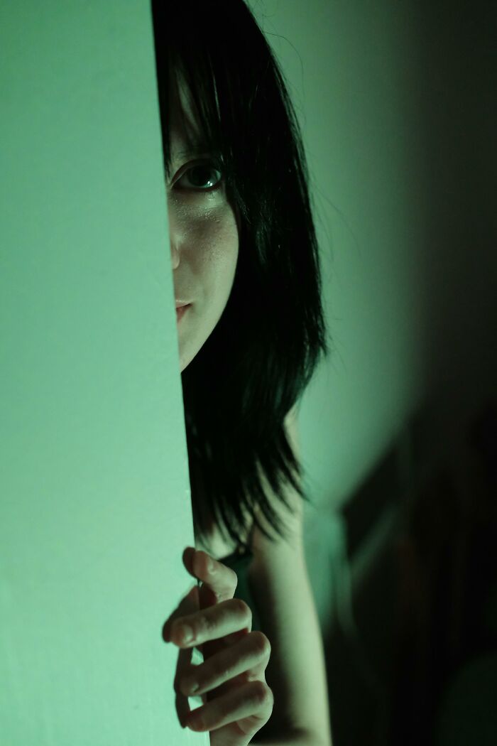 Person peeking from behind a wall in dim green light, evoking mysterious neighbor tales in a wooded setting.