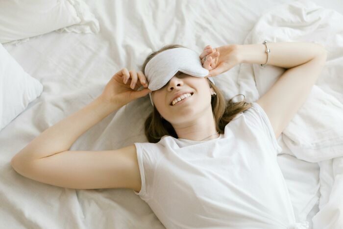 A smiling woman wearing a sleep mask lying on a bed, illustrating people share things that improved life. - 22