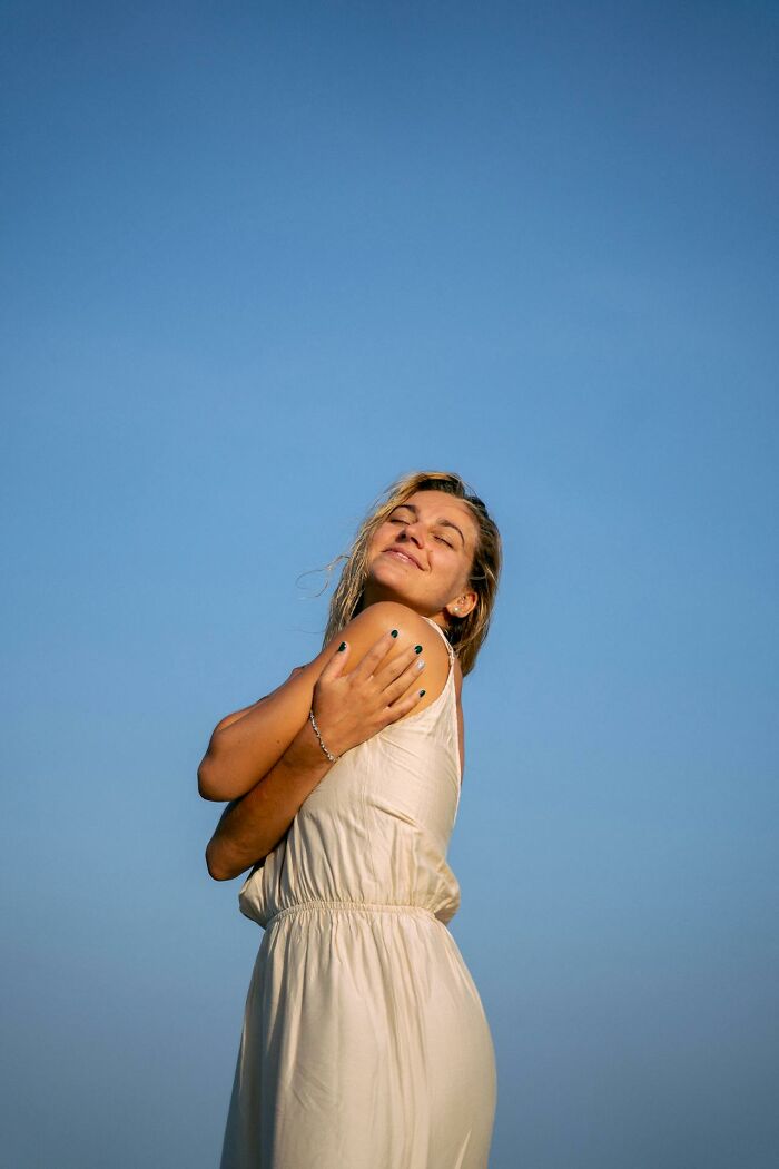 Young woman in a light dress outdoors smiling with eyes closed, embracing herself, promoting stay hydrated wellness.