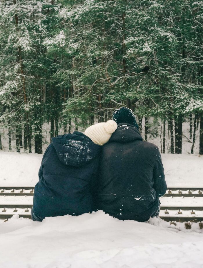 Two people dressed in winter clothing sit on snow near train tracks with a forest in the background, survival tips theme.