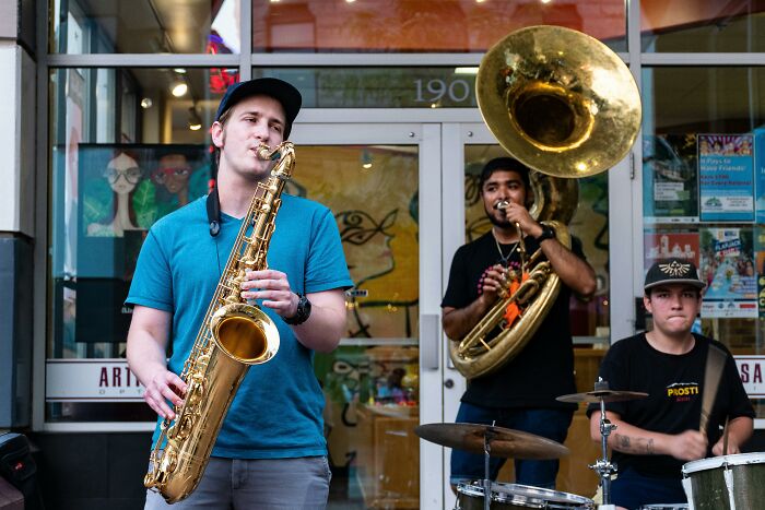 Three street musicians playing saxophone, tuba, and drums, showcasing creative student names in a lively setting.