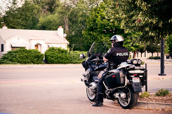 Police officer sitting on a motorcycle wearing helmet and uniform near a roadside tree in a suburban area, survival tips concept.