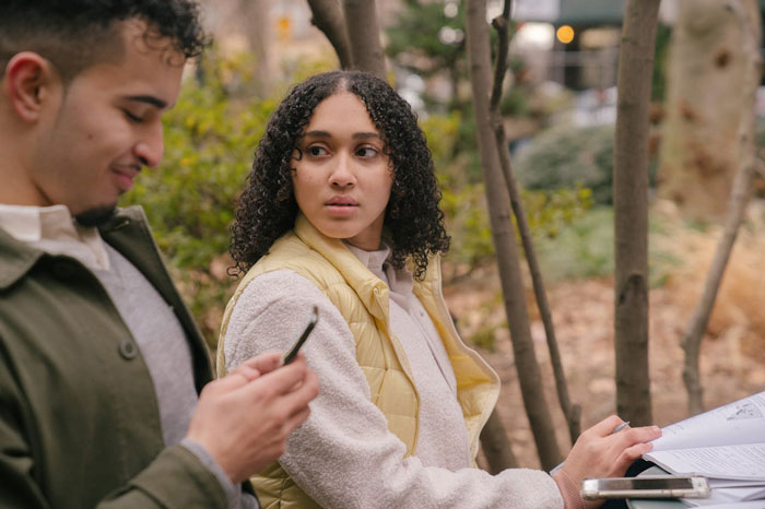 Woman looks suspiciously at her husband using phone while sitting outdoors, depicting jealous wife spying husband work scenario.