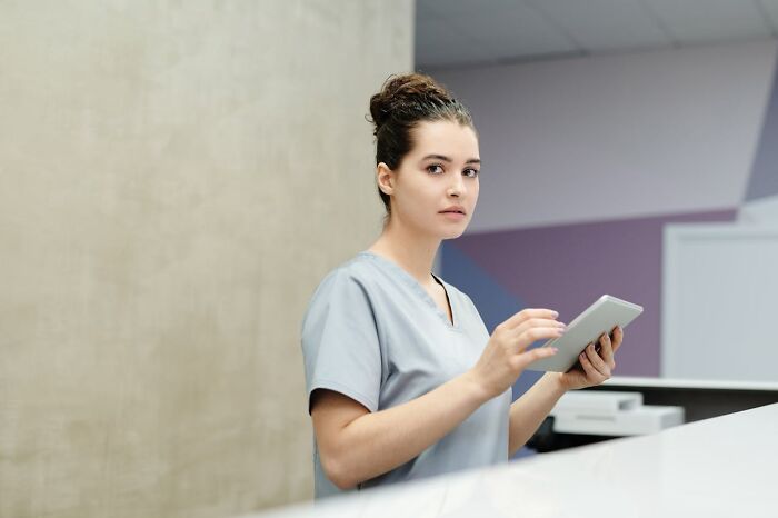 Female nurse in scrubs holding a tablet, standing at a hospital reception with a focused expression on her face