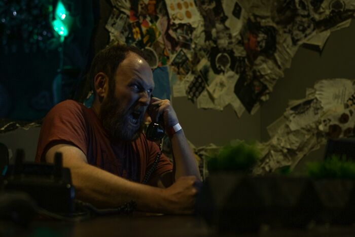 Man angrily talking on the phone in a dim room, surrounded by notes and papers, evoking neighbor tales in the woods.