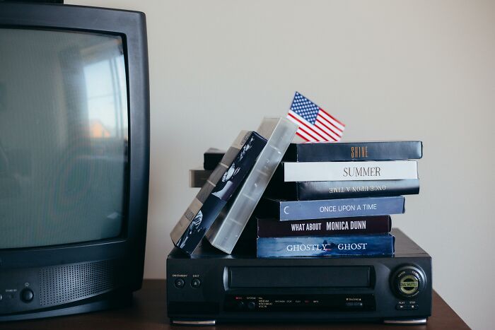 Stack of classic movie DVDs and VHS tapes with an American flag on a VCR next to a vintage television set.