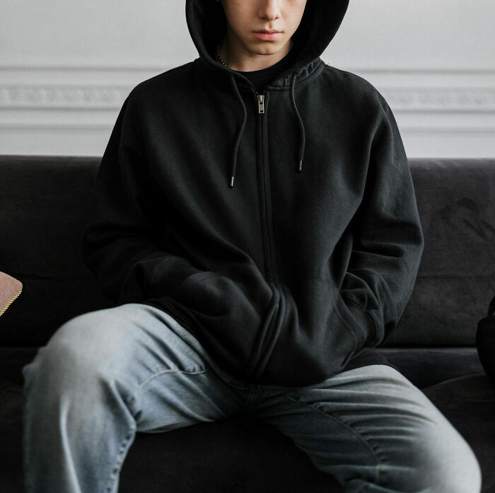 Teenager wearing a black hoodie and jeans sitting on a couch, conveying a disturbing vibe related to overheard statements.