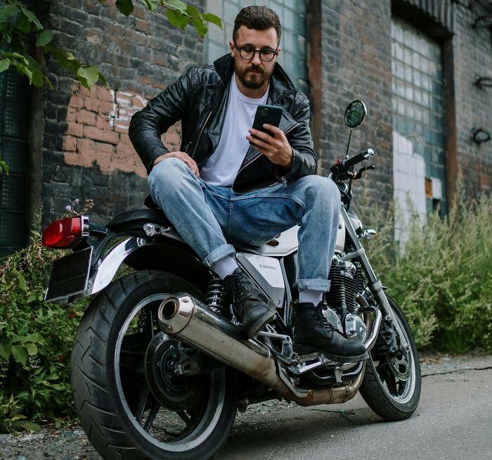 Man with glasses sitting on a motorcycle near graffiti wall, using phone, illustrating truckers share craziest things seen concept. - 10