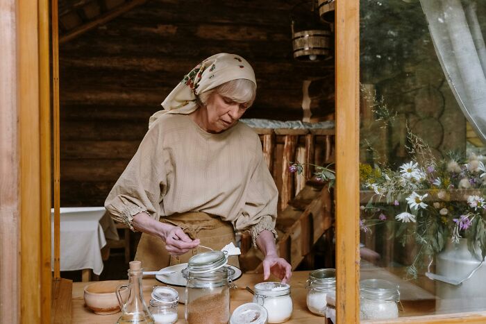 Elderly woman in traditional clothing preparing food in a rustic kitchen, surrounded by jars and family lore atmosphere.