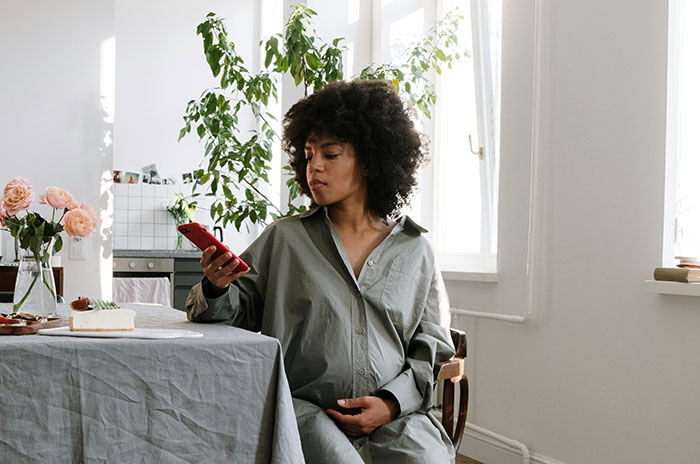 Pregnant woman sitting at table, looking at phone, concerned about neighbor lurking around her house repeatedly. - 31