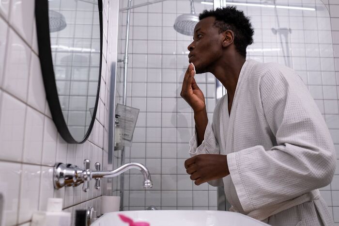 Man in a white robe examining his face in a bathroom mirror, reflecting on cheating red flags in a relationship. - 20