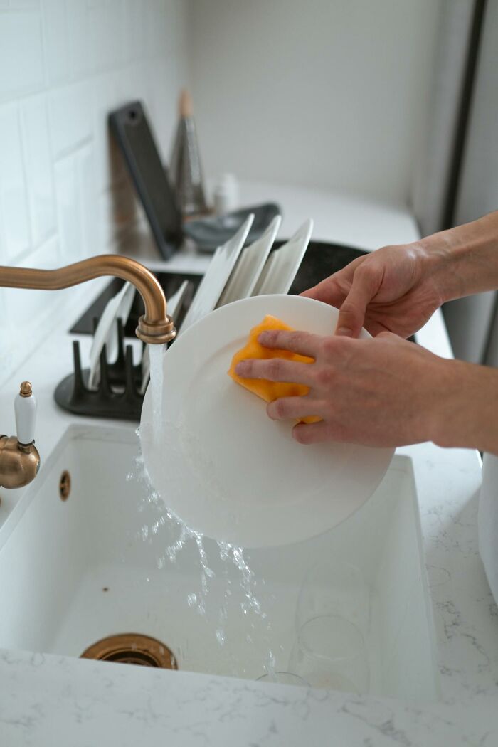 Hands washing dishes under running water in a kitchen sink, emphasizing the importance to stay hydrated throughout the day.