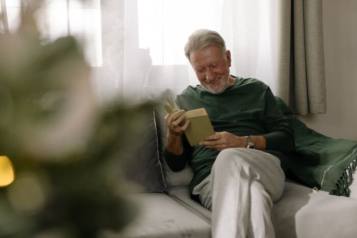 Older man sitting on a couch at home, reading a book and reflecting on life after divorce. - 20